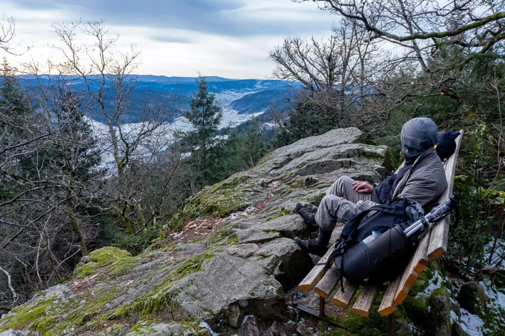 Wanderer sitzt auf Bank, die auf einem Felsen steht und schaut in die Ferne