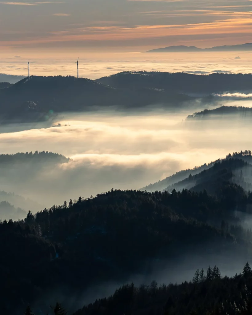 Aussicht von oben auf Nebel zwischen Bergen und Windrädern im Hintergrund